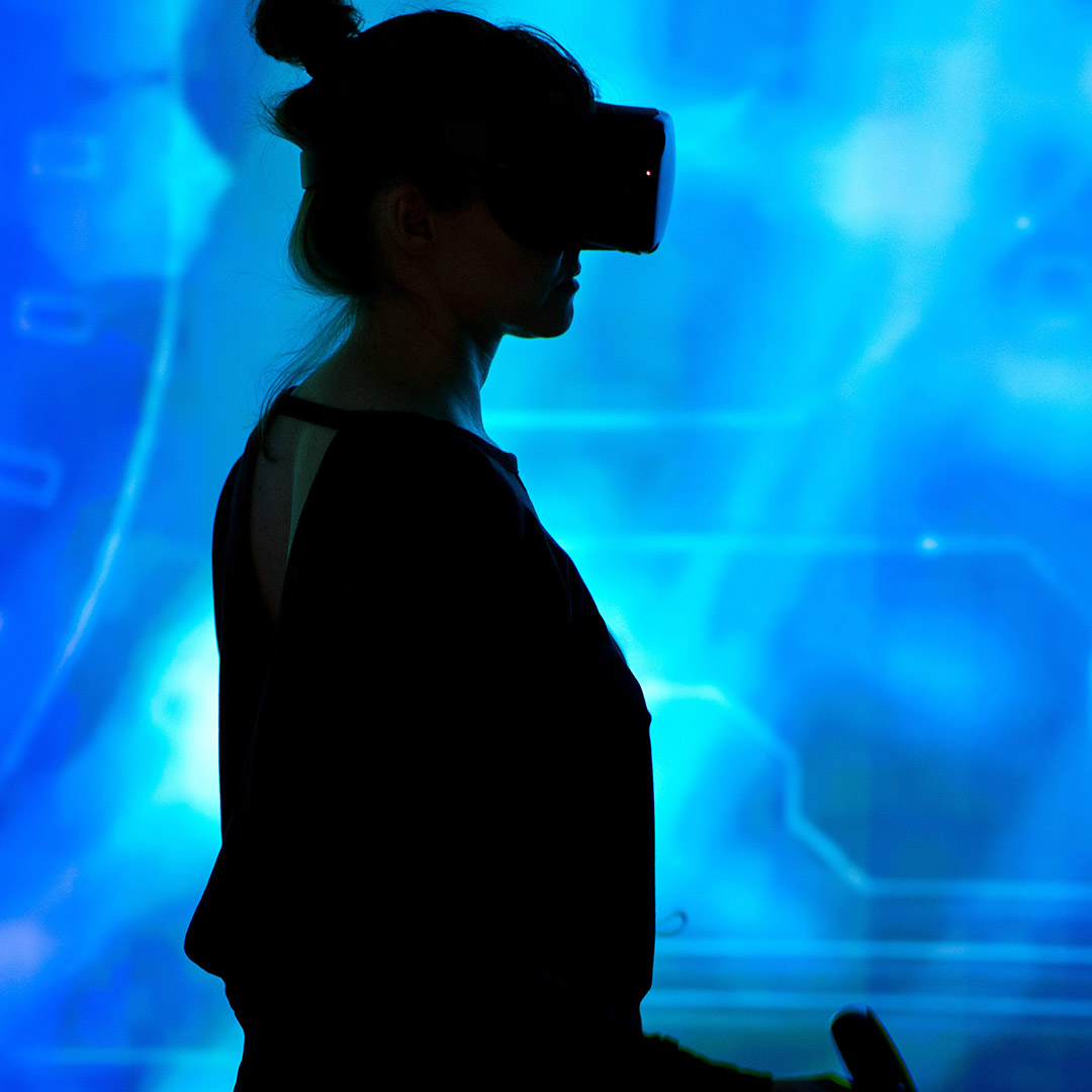 Photo of a woman with VR glasses standing sideways against a blue background.