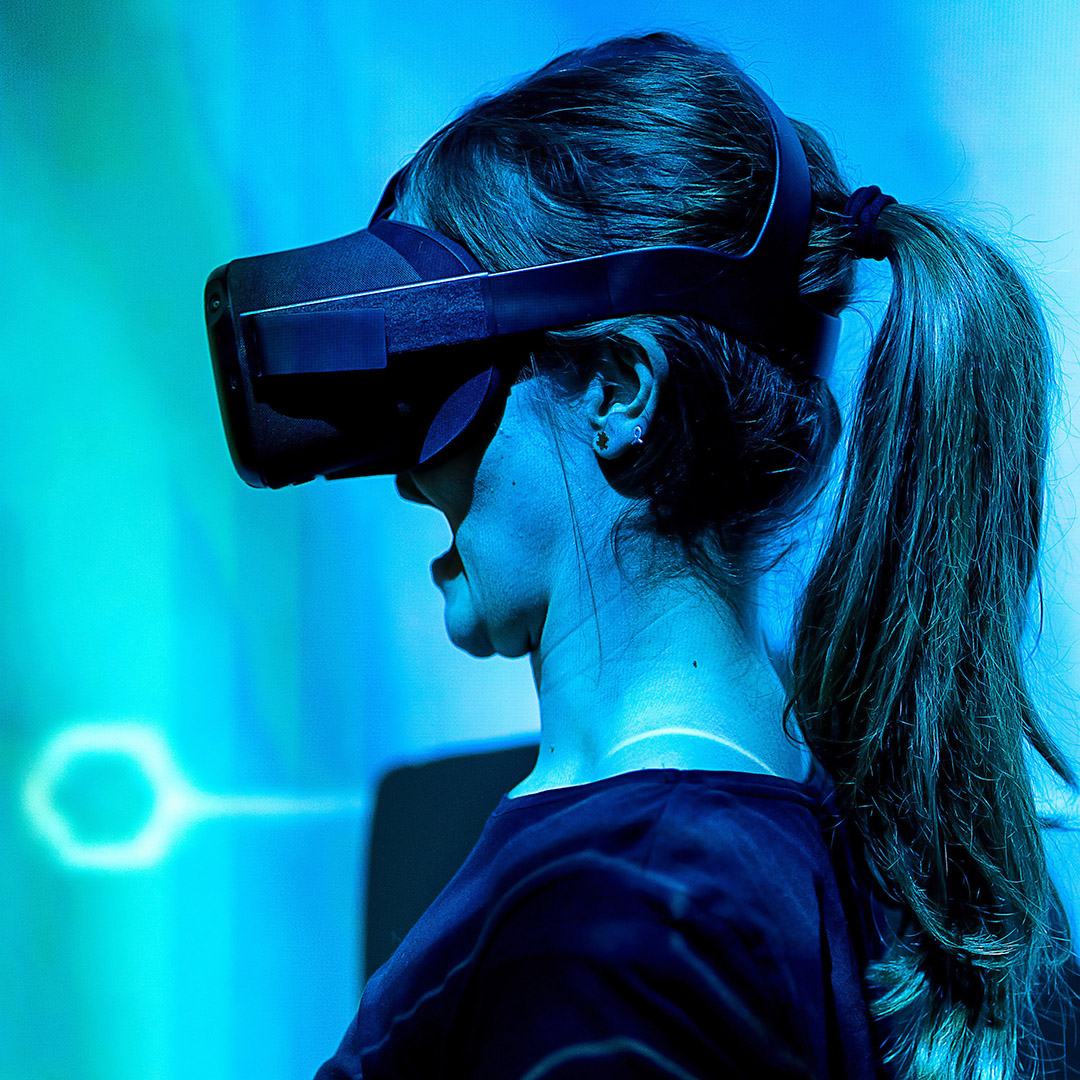 Photo of a bust of a woman standing sideways, wearing VR glasses, with her mouth open. Picture taken in blue light background.