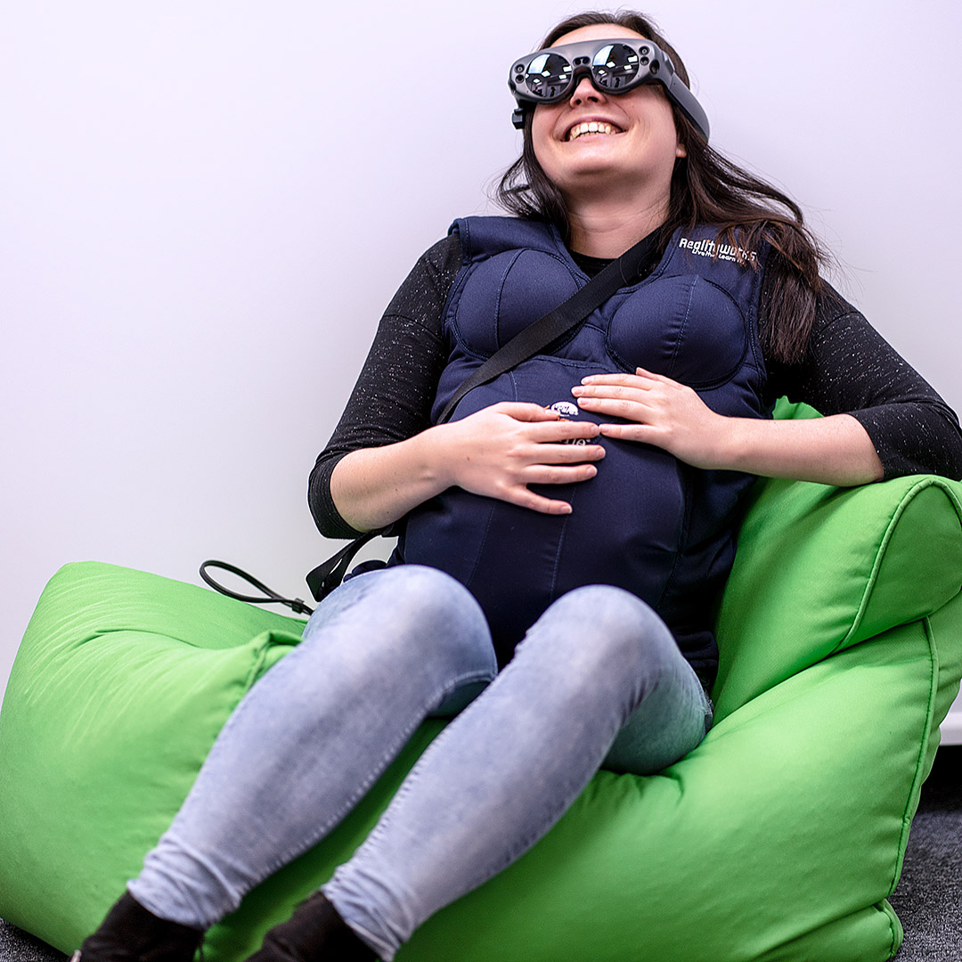 Photo of a smiling woman sitting on a green pouf, wearing a suit simulating the state of pregnancy (enhancing her breasts and a large belly), wearing VR glasses. A photo is taken on a light background.
