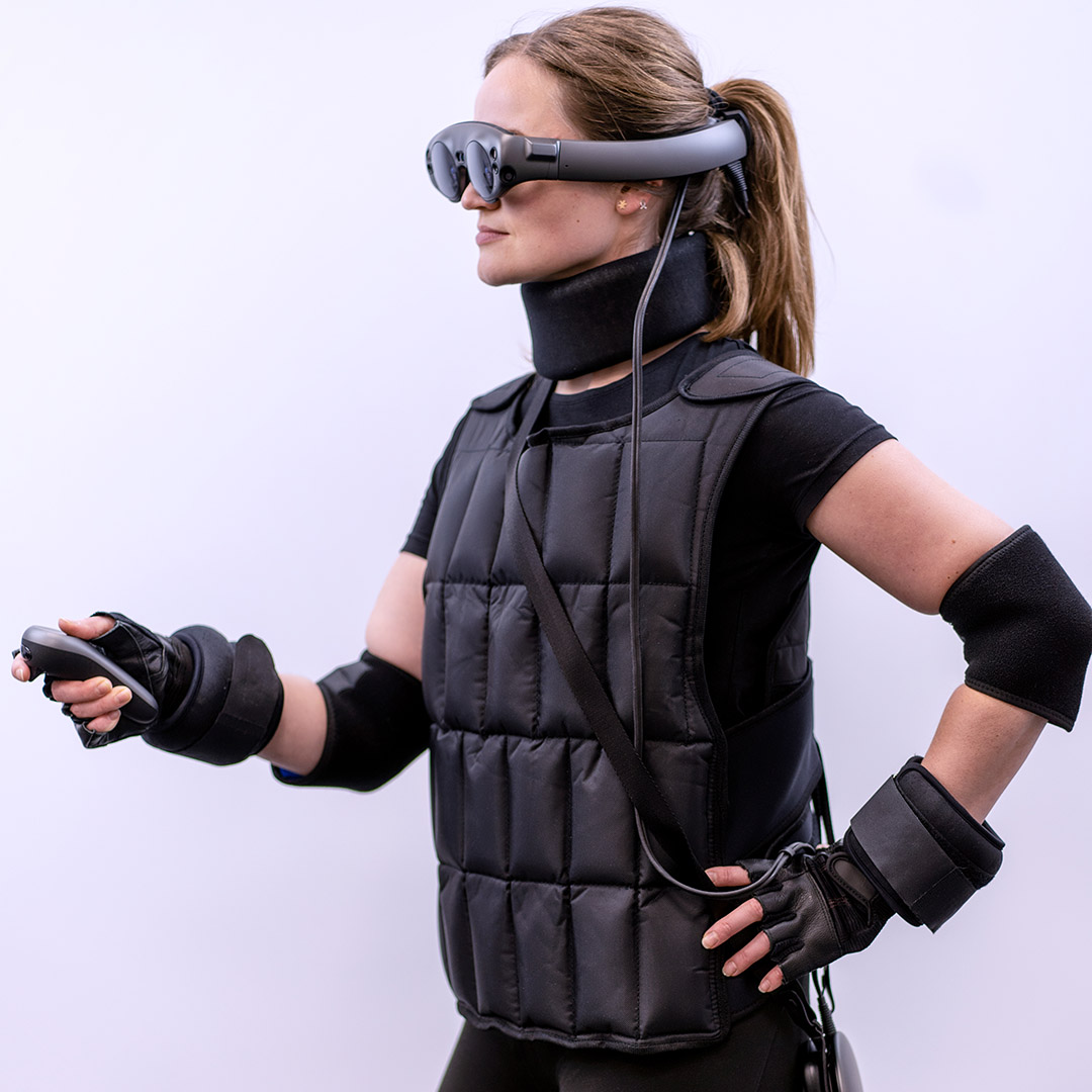 A photo of a woman standing sideways, with her left hand under her side, and her right outstretched hand operating the VR controller. The woman is wearing VR glasses and a black geriatric suit, which consists of a vest, elbow bands, wrists and neck, as well as gloves. Picture taken on a light background.